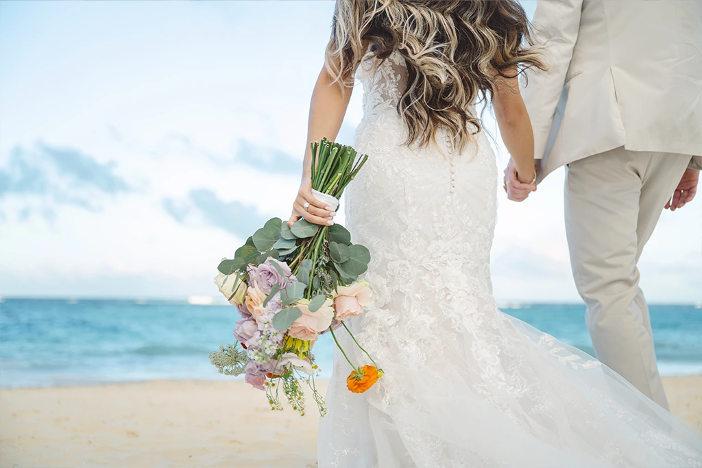 A couple walking on the beach and holding hands, bride with the flowers in her other hand
