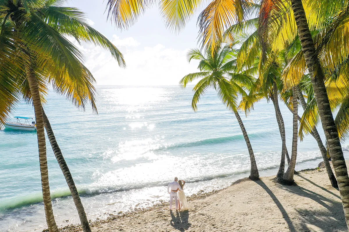 Aerial drone view of a couple on a palm-lined Punta Cana beach beside turquoise water and a boat