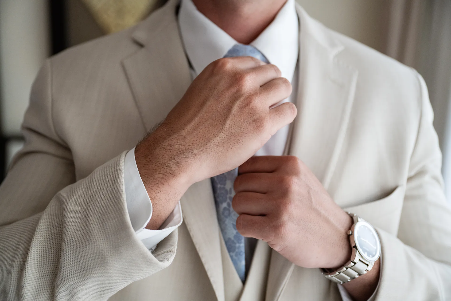 Groom adjusting a light blue floral tie on his beige three-piece beach wedding suit