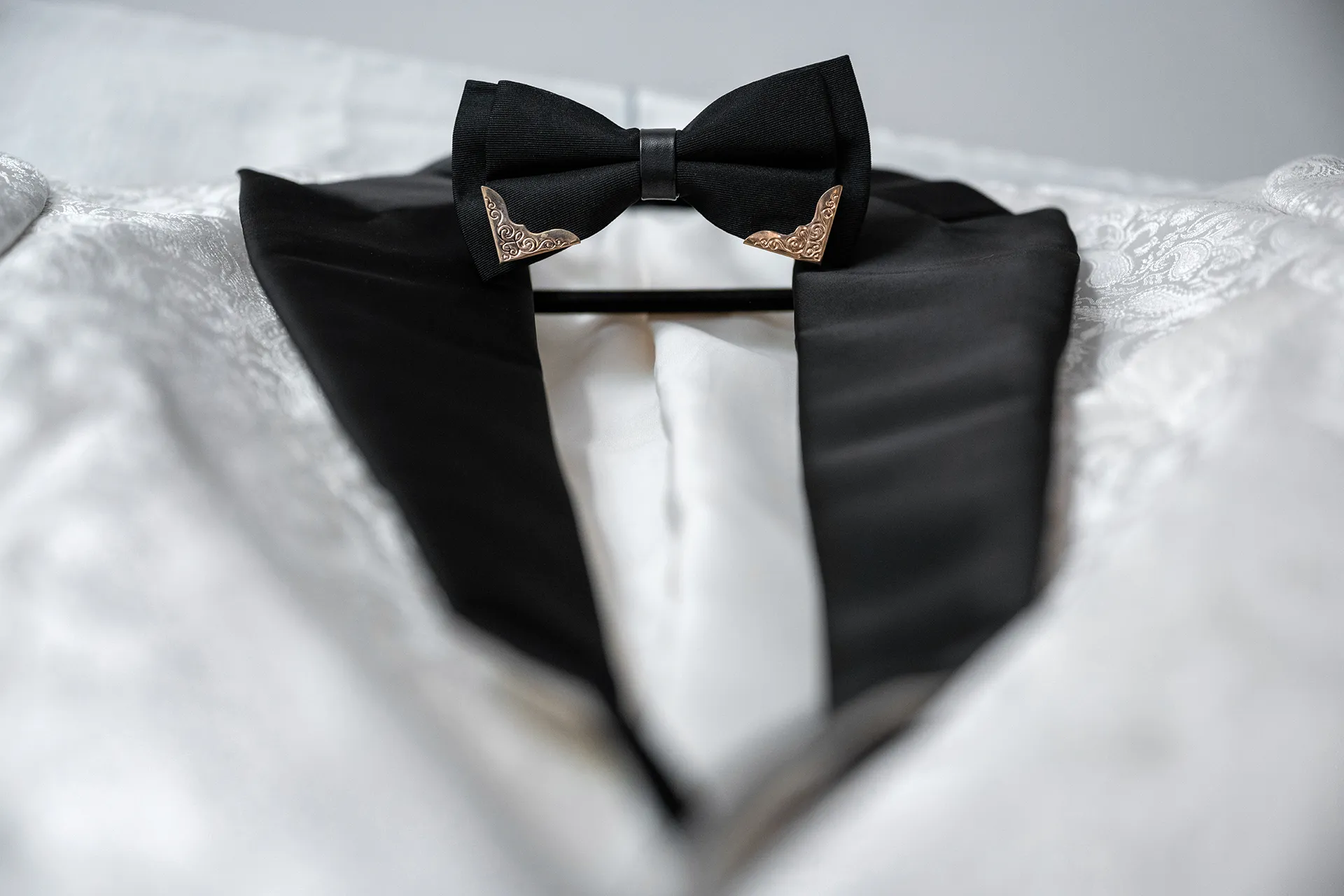 black-bow-tie-groom-wedding-details Black bow tie with ornate gold tips resting on a groom's white jacquard dinner jacket