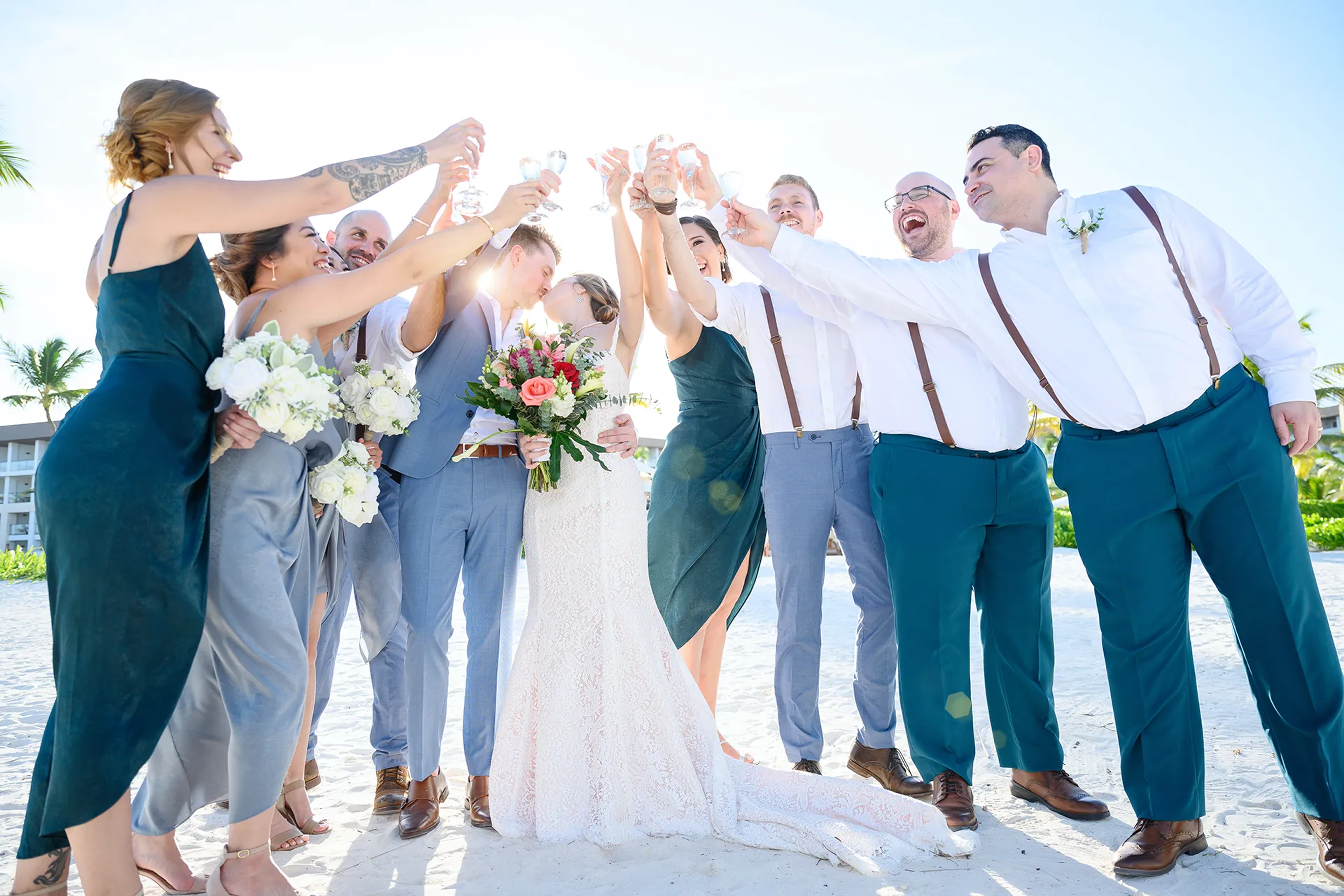 Wedding party toasting on the sand in teal bridesmaid dresses, light suits and suspenders at a Punta Cana beach wedding