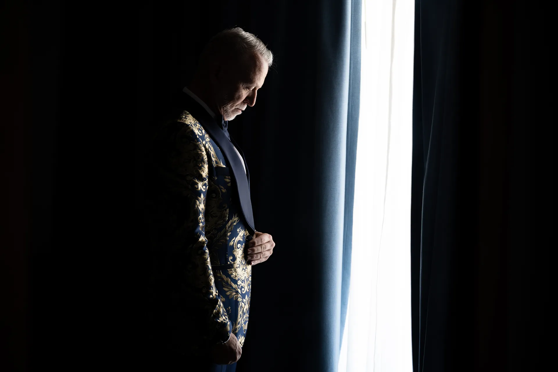 Groom buttoning a navy and gold brocade dinner jacket by the window while getting ready