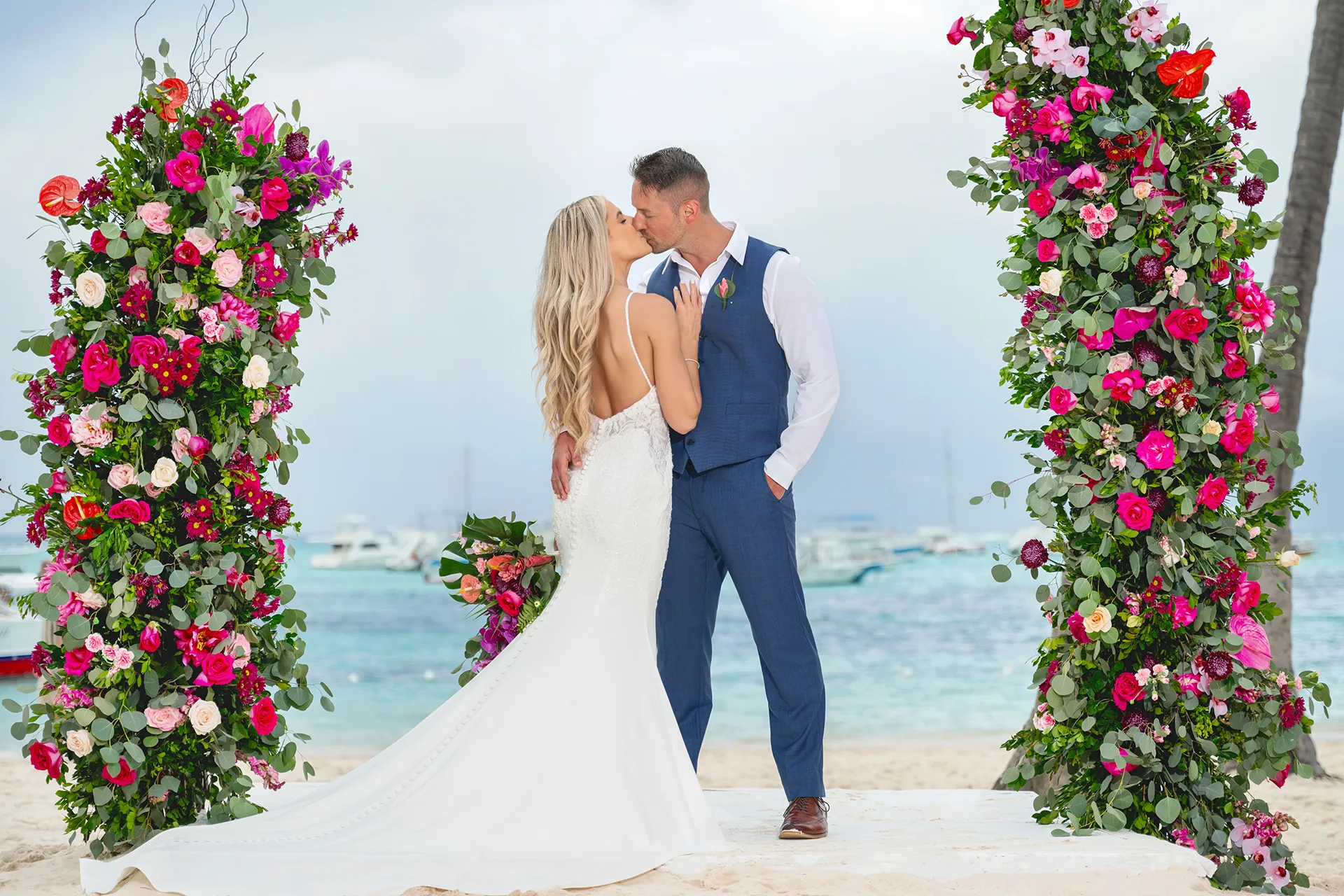 Bride in a fitted lace gown and groom in a blue vest and trousers kissing under a floral arch on a Punta Cana beach