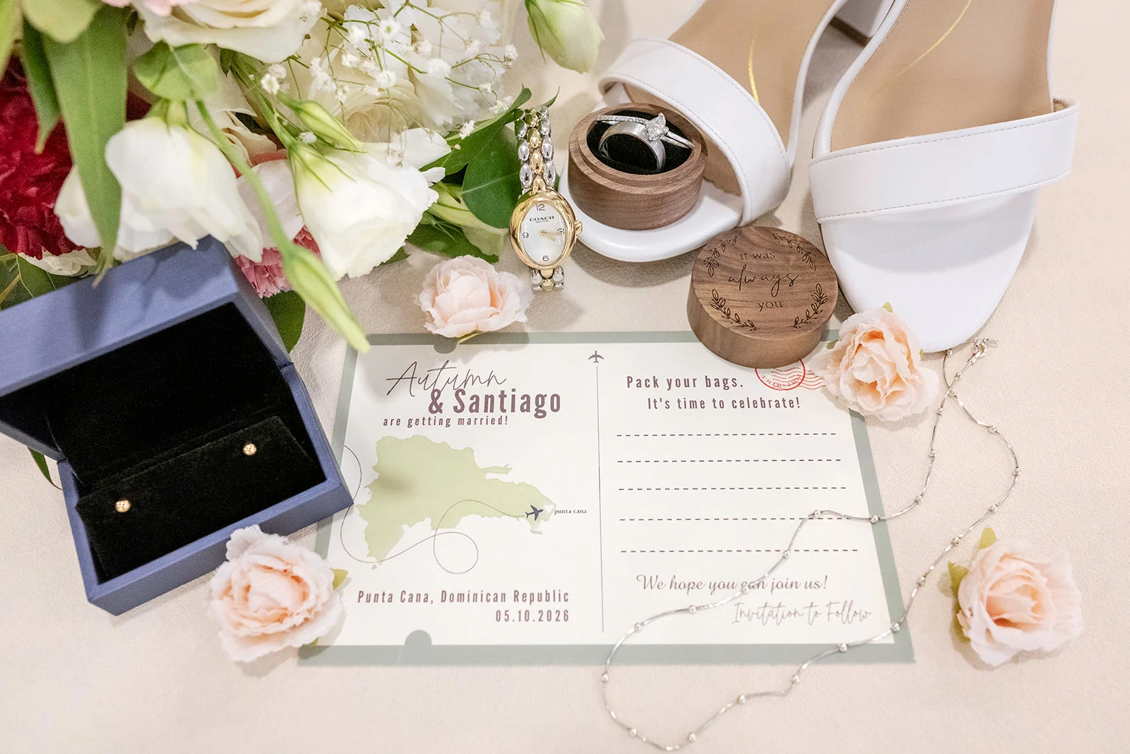 dreams-flora-bridal-wedding-details Wedding details flat lay with a travel-themed save-the-date postcard, an engraved wooden ring box, white sandals, a watch and blush roses