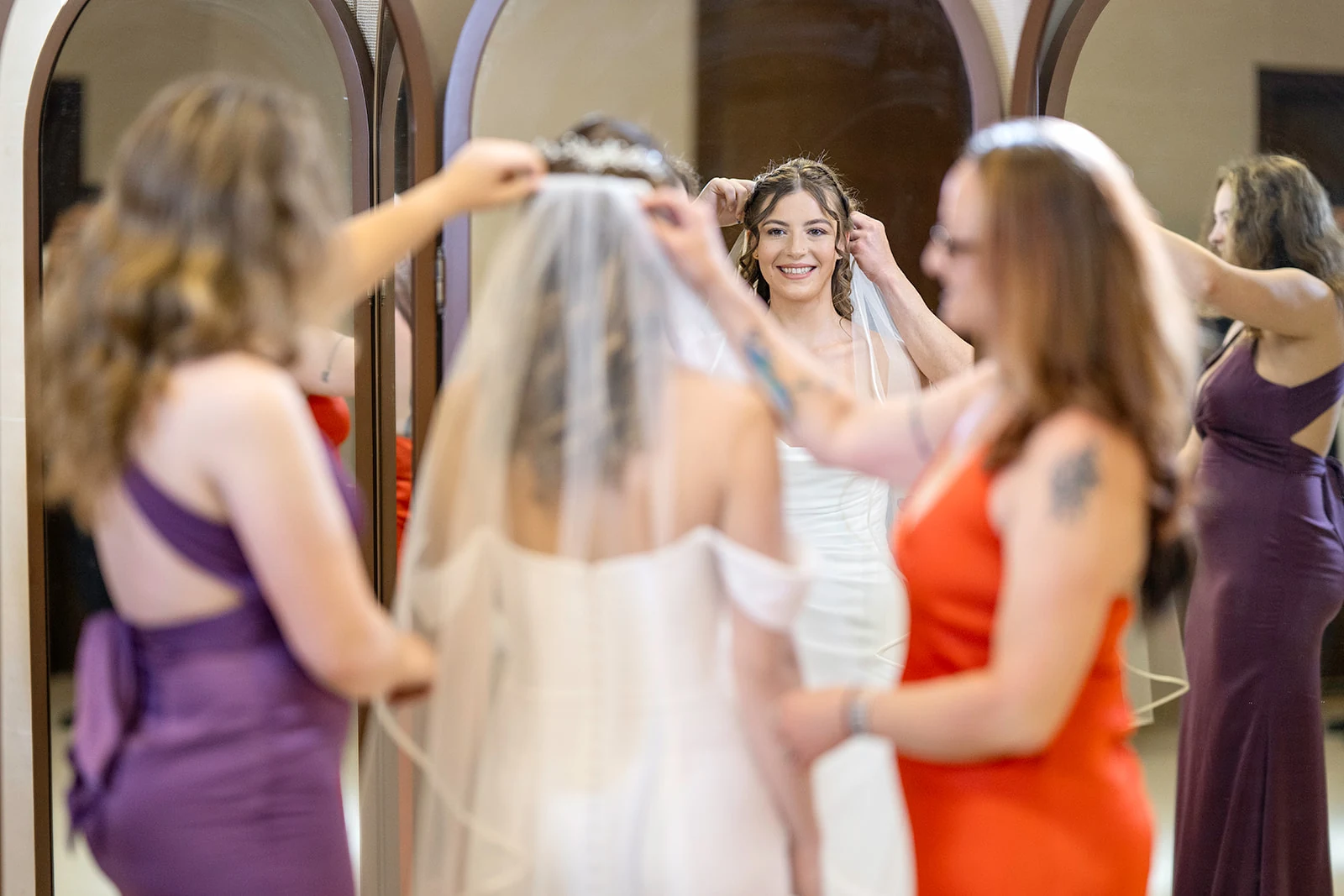 dreams-flora-bride-getting-ready-with-bridesmaids Bride smiling at her reflection as bridesmaids in purple and orange dresses place her veil