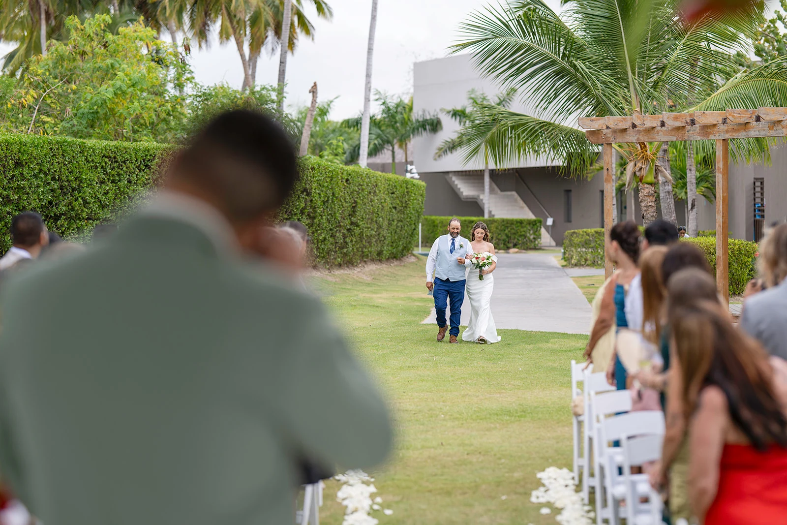 Bride escorted down the grass aisle toward the garden gazebo at Dreams Flora as the groom waits in the foreground