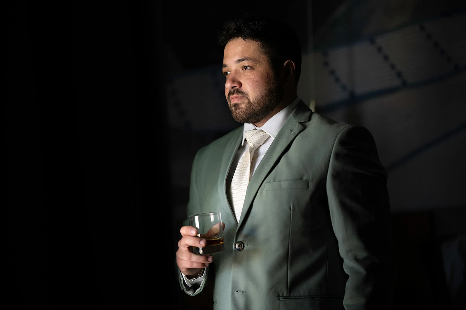 Groom in a sage green suit holding a glass of whiskey in dramatic window light before the ceremony
