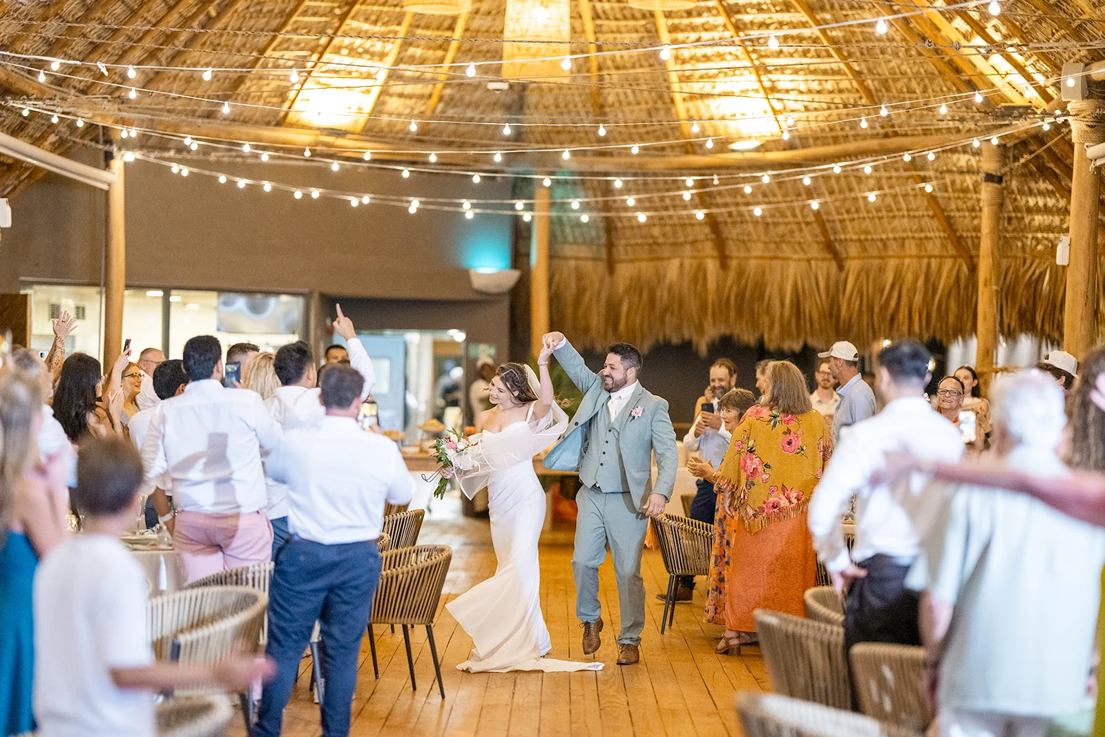 dreams-flora-open-air-wedding-reception-entrance Bride and groom making their grand entrance under a thatched roof and string lights as guests cheer