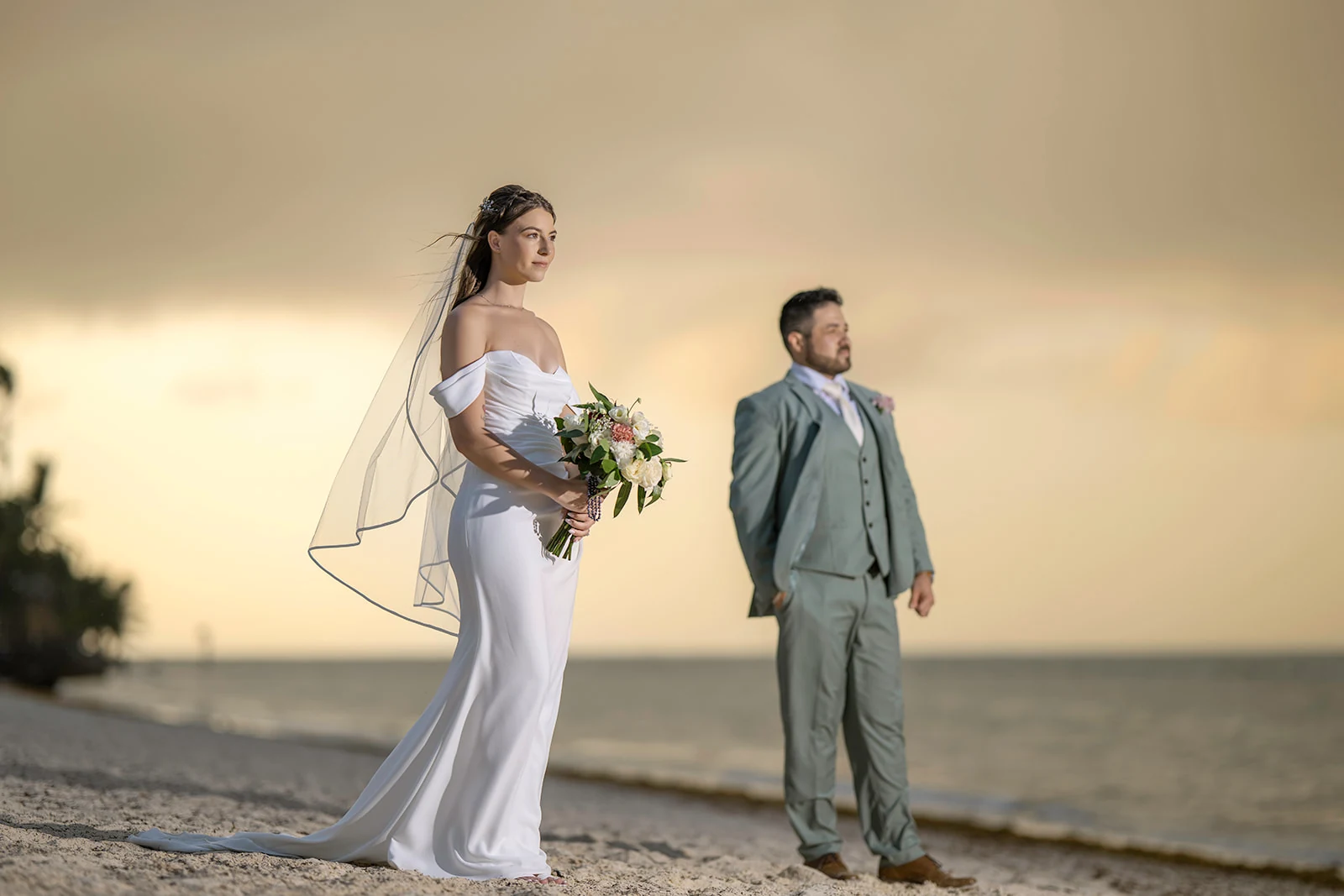 dreams-flora-punta-cana-beach-wedding-portrait Bride holding her bouquet on the beach at sunset with the groom standing behind her in soft focus