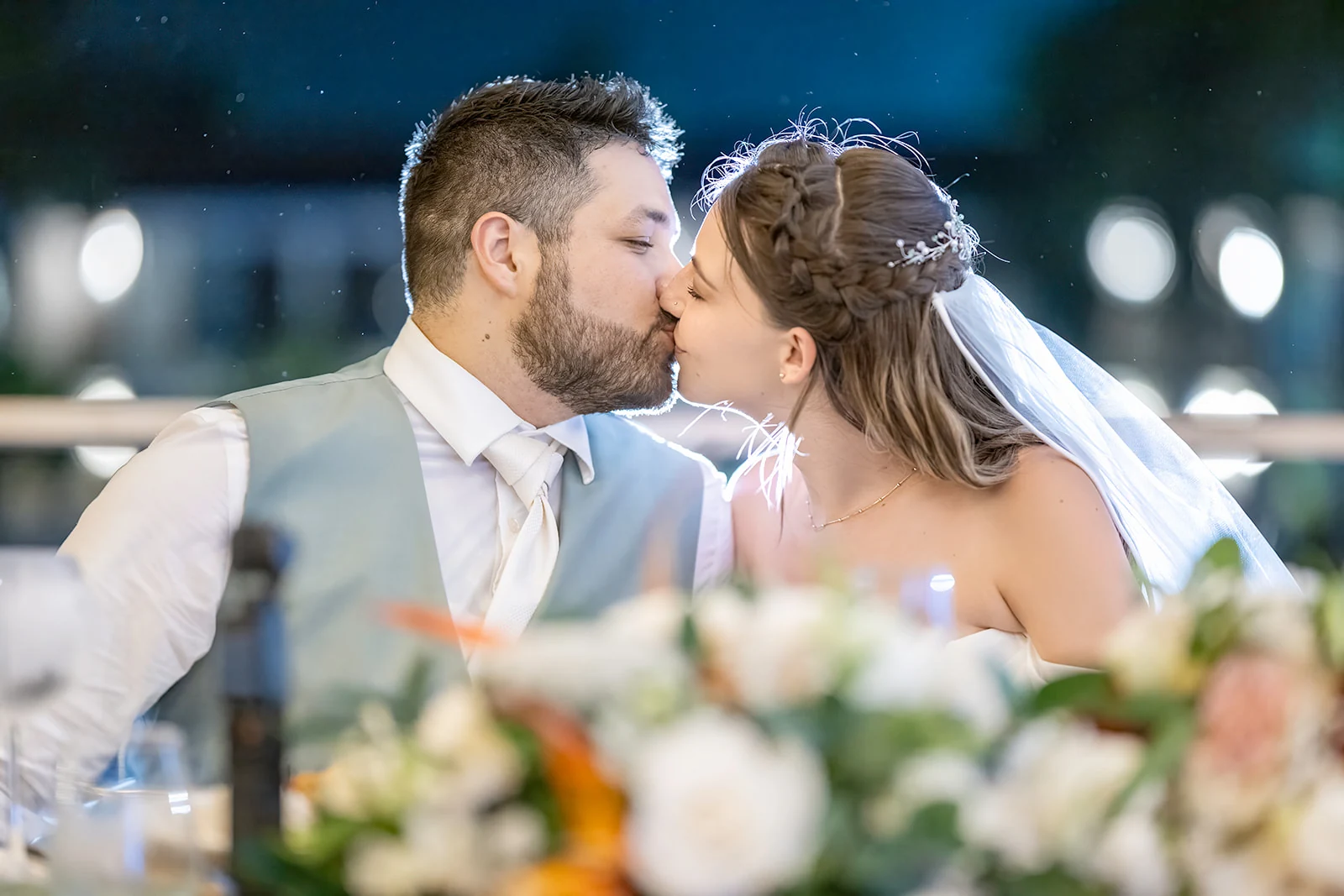 dreams-flora-wedding-couple-evening-portrait Bride and groom kissing at their reception table behind a soft blur of white and peach flowers